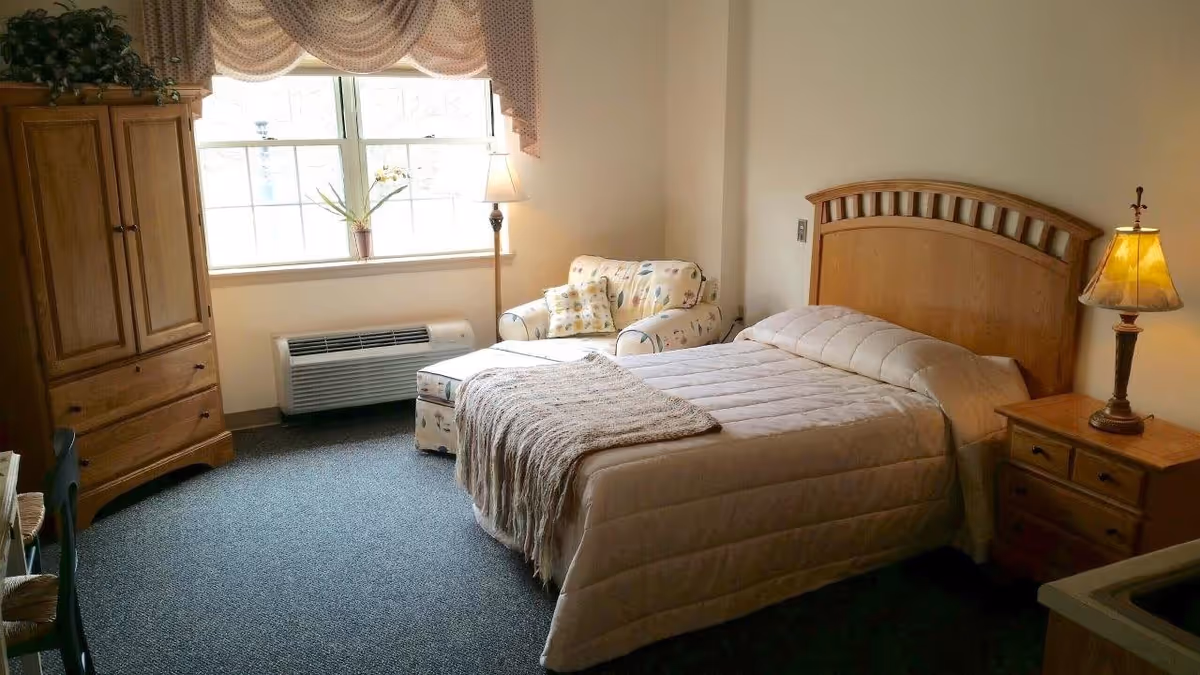 A cozy bedroom in Woodview Estates featuring a wooden bed with a beige comforter and a matching wooden nightstand with a lamp. There is a cushioned armchair with a floral pattern near a window with draped curtains. A wooden armoire and a small table with chairs are also visible, along with a floor lamp and a heating unit beneath the window.