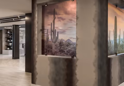 Interior view of a modern hallway with a large framed photograph of a desert landscape featuring tall cacti mounted on a wall. The hallway has light wood flooring and dark structural columns, with shelves and additional decor visible in the background.