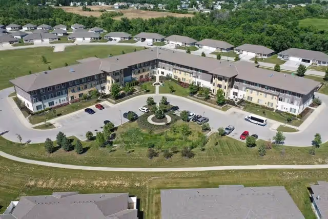 Aerial view of The Piper Assisted Living and Memory Care facility showing a large U-shaped building with a parking lot in front, surrounded by greenery and residential houses in the background.