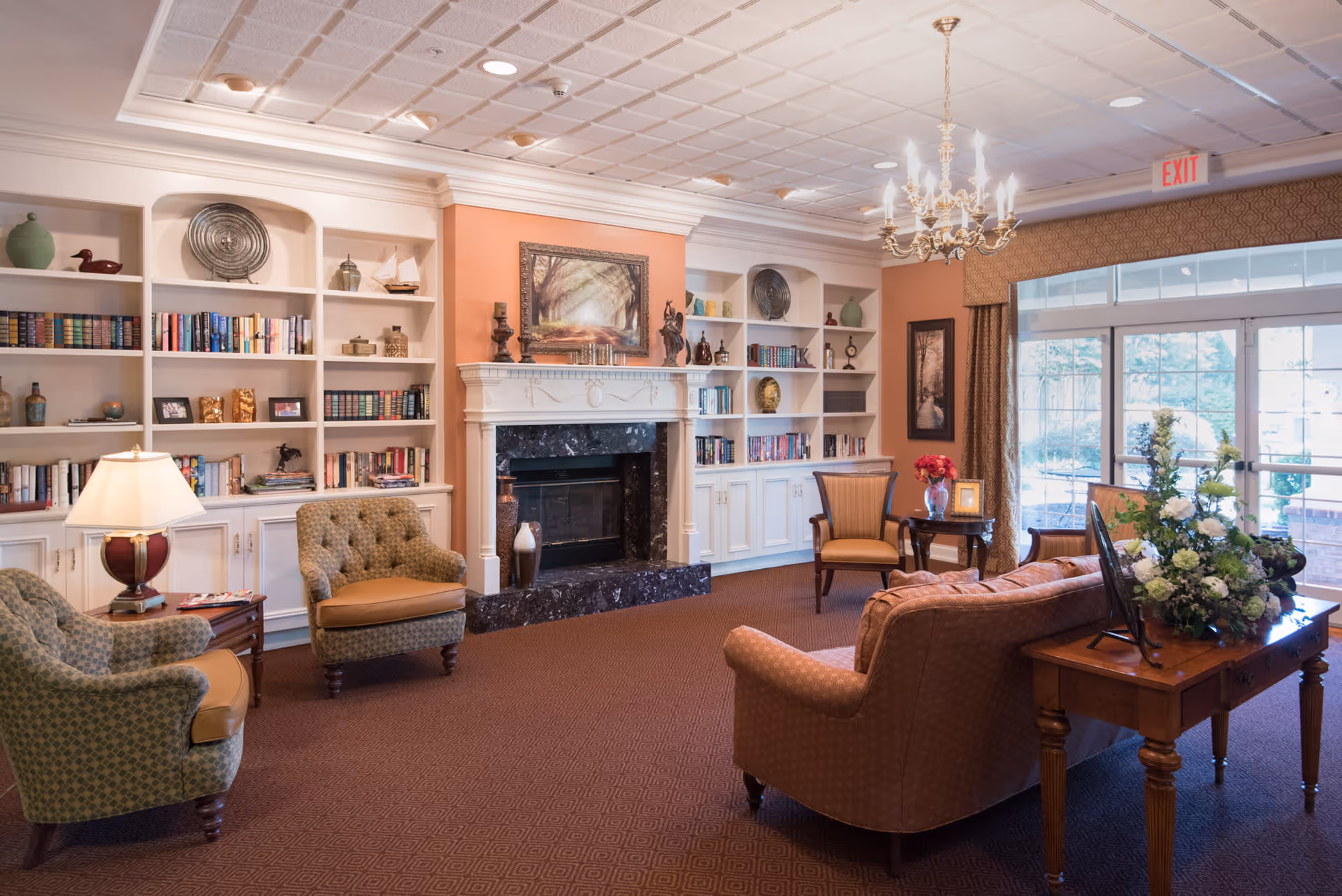 A cozy living room area with a fireplace centered between built-in white bookshelves filled with books and decorative items. The room features upholstered armchairs and a sofa arranged around a wooden table with a floral arrangement. A chandelier hangs from the ceiling, and large windows with curtains allow natural light to fill the space.