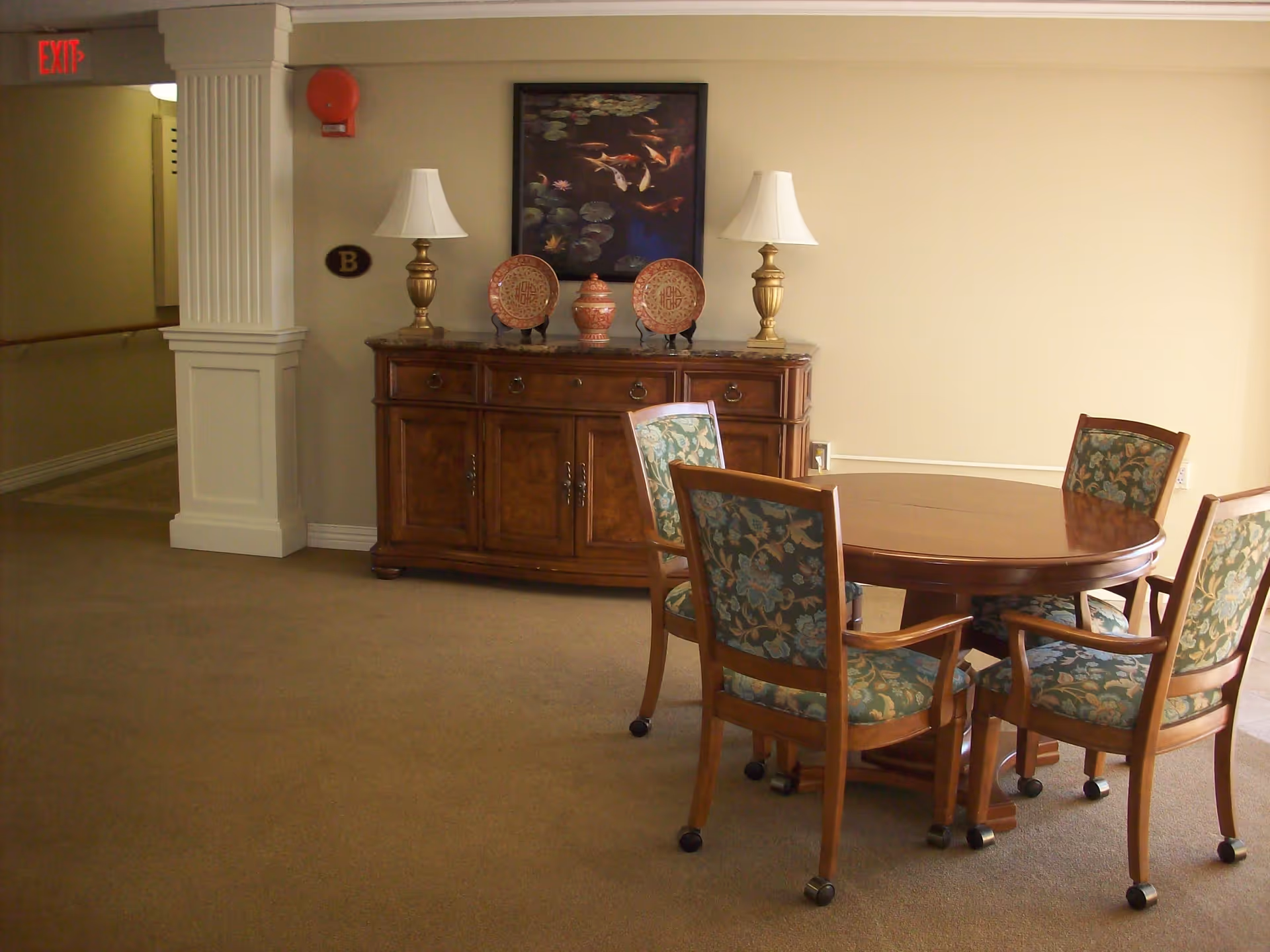 Round wooden dining table with four floral-upholstered chairs and a wooden sideboard with lamps and decorative plates in a senior living interior.