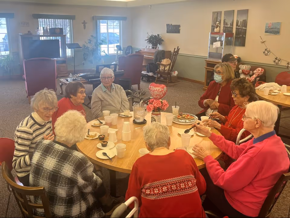 A group of elderly residents seated around a round table in a senior living common room sharing snacks and drinks.