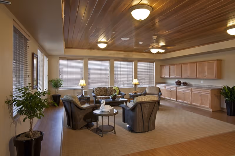 A spacious living room area with a wooden ceiling, multiple ceiling lights, and large windows with blinds. The room features a seating arrangement with a sofa and four armchairs around a coffee table, two side tables with lamps, and a long wooden cabinet along one wall. There are also several potted plants placed around the room.