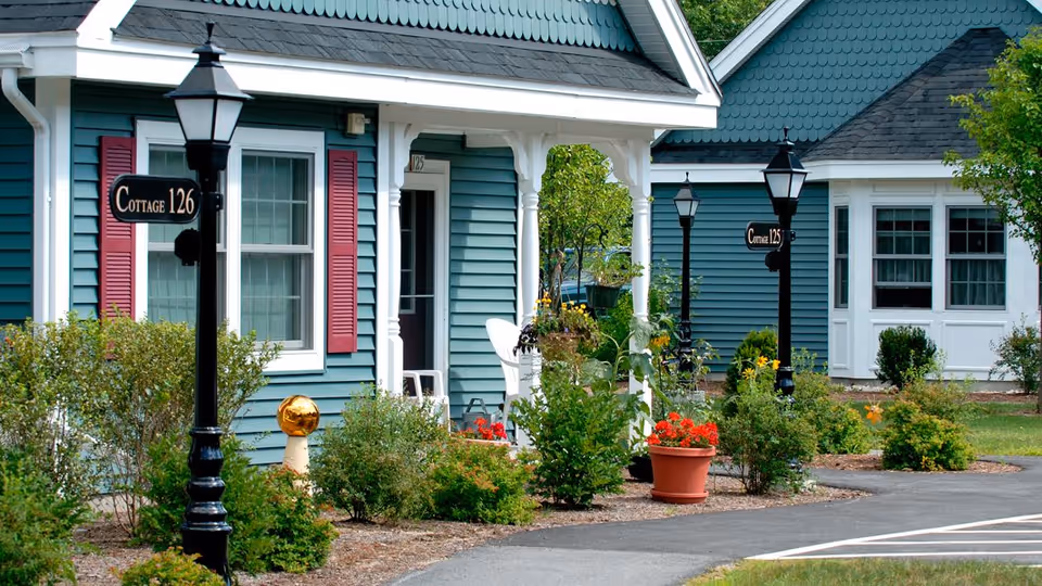Exterior view of blue cottages with white trim and red shutters at a senior living facility. There are black lamp posts with signs indicating Cottage 126 and Cottage 125. The area is landscaped with green bushes, potted flowers, and a paved walkway.