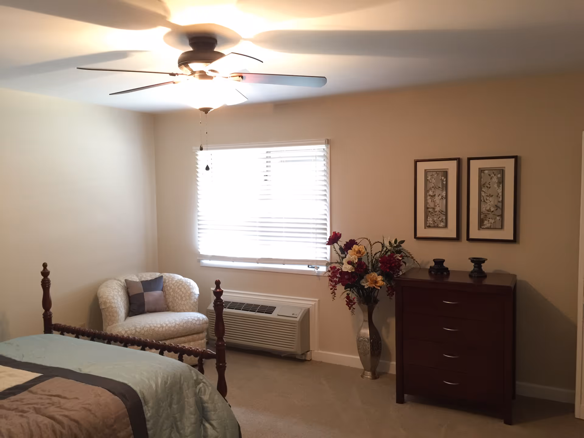 A cozy bedroom with a wooden bed frame partially visible, a cushioned armchair with a pillow near a window with blinds, a ceiling fan with lights, a wooden dresser with decorative items on top, and a tall vase with artificial flowers next to the dresser. Two framed pictures hang on the wall above the dresser.