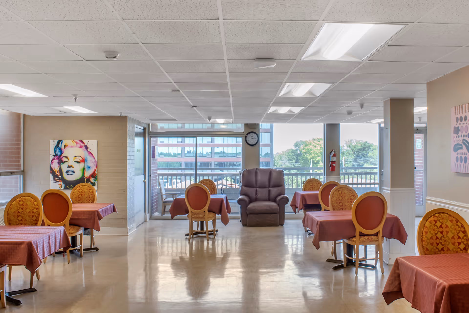 Dining room with multiple small tables covered in red tablecloths, patterned chairs, a central recliner, and large windows at the far end.