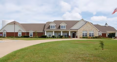 Single-story senior living facility building with a combination of brick and stone exterior, multiple dormer windows on the roof, a covered entrance, and a well-maintained lawn in front under a partly cloudy sky.