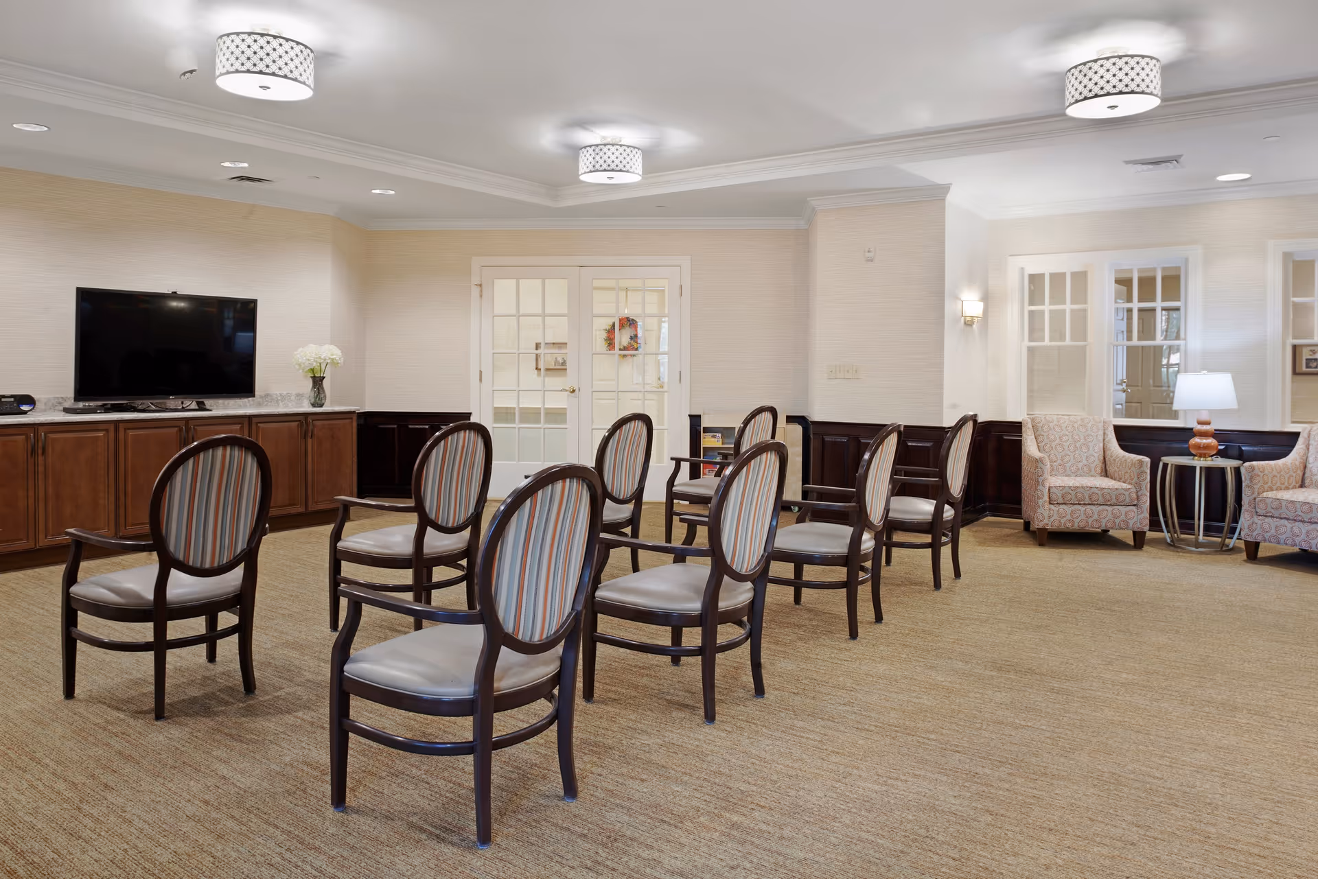 Bright common room with rows of wooden chairs facing a wall-mounted TV and two upholstered armchairs by a side table.