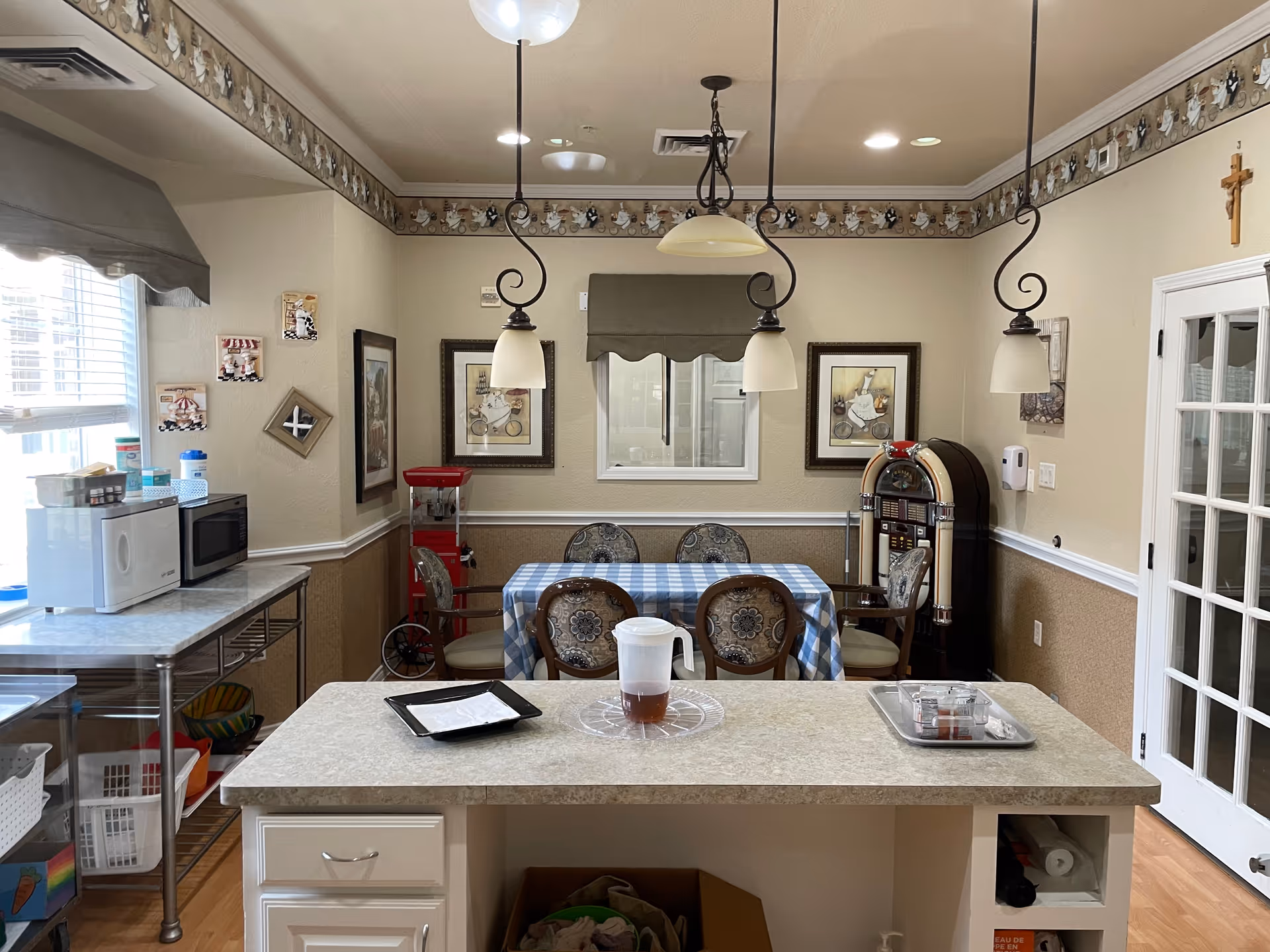 A cozy dining area in Magnolia Assisted Living and Memory Care featuring a table covered with a blue and white checkered tablecloth surrounded by four chairs. The room has beige walls with decorative border trim near the ceiling, three hanging pendant lights, framed artwork on the walls, a vintage-style jukebox in the corner, and a popcorn machine. A kitchen island with a pitcher and trays is in the foreground, and a microwave and other kitchen items are on a metal rack to the left. A window with a valance and a glass-paneled door are also visible.