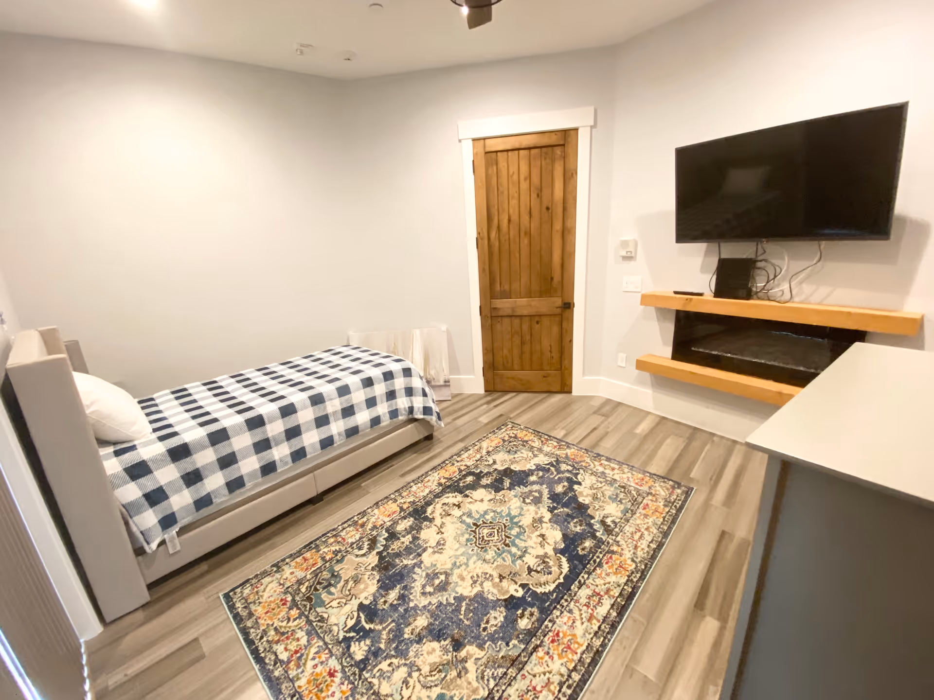 A small, neatly arranged bedroom with a single bed covered in a blue and white checkered blanket. There is a colorful patterned rug on the wooden floor. A wooden door is centered on the far wall. A flat-screen TV is mounted on the wall above a modern electric fireplace with wooden shelves. A gray dresser is partially visible on the right side.