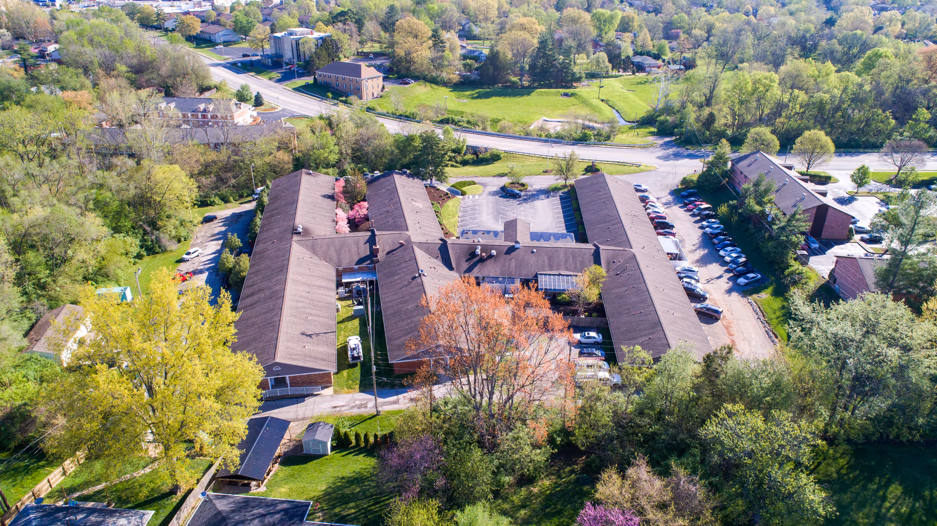 Aerial view of Hillcreek Rehabilitation and Care facility surrounded by trees and greenery, showing multiple connected buildings with brown roofs, parking lots with cars, and nearby roads.