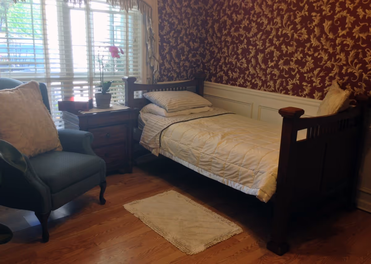A cozy bedroom with a wooden single bed, armchair, nightstand, and ornate wallpaper.