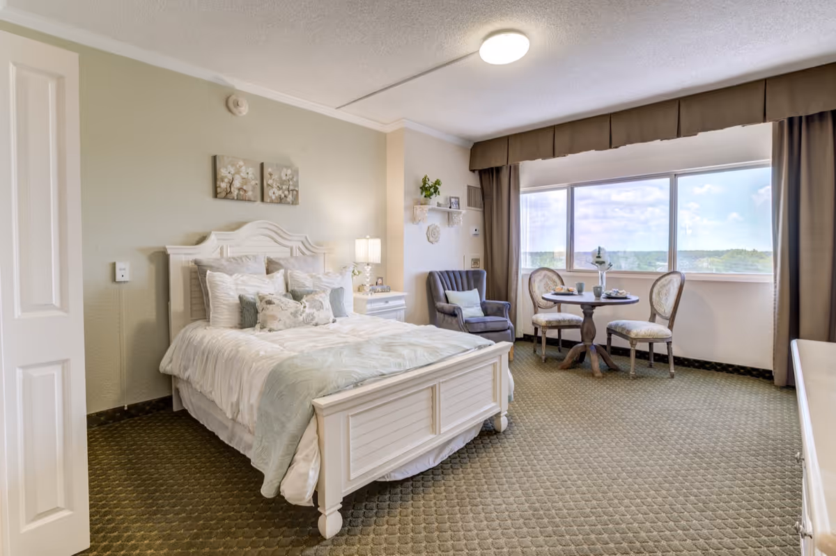 A bright and cozy bedroom in a senior living facility featuring a white wooden bed with multiple pillows and a light blue throw blanket. Next to the bed is a white nightstand with a lamp and decorative items. A comfortable armchair is placed near a large window with brown curtains, and a small round table with two upholstered chairs is set by the window, offering a scenic view outside.