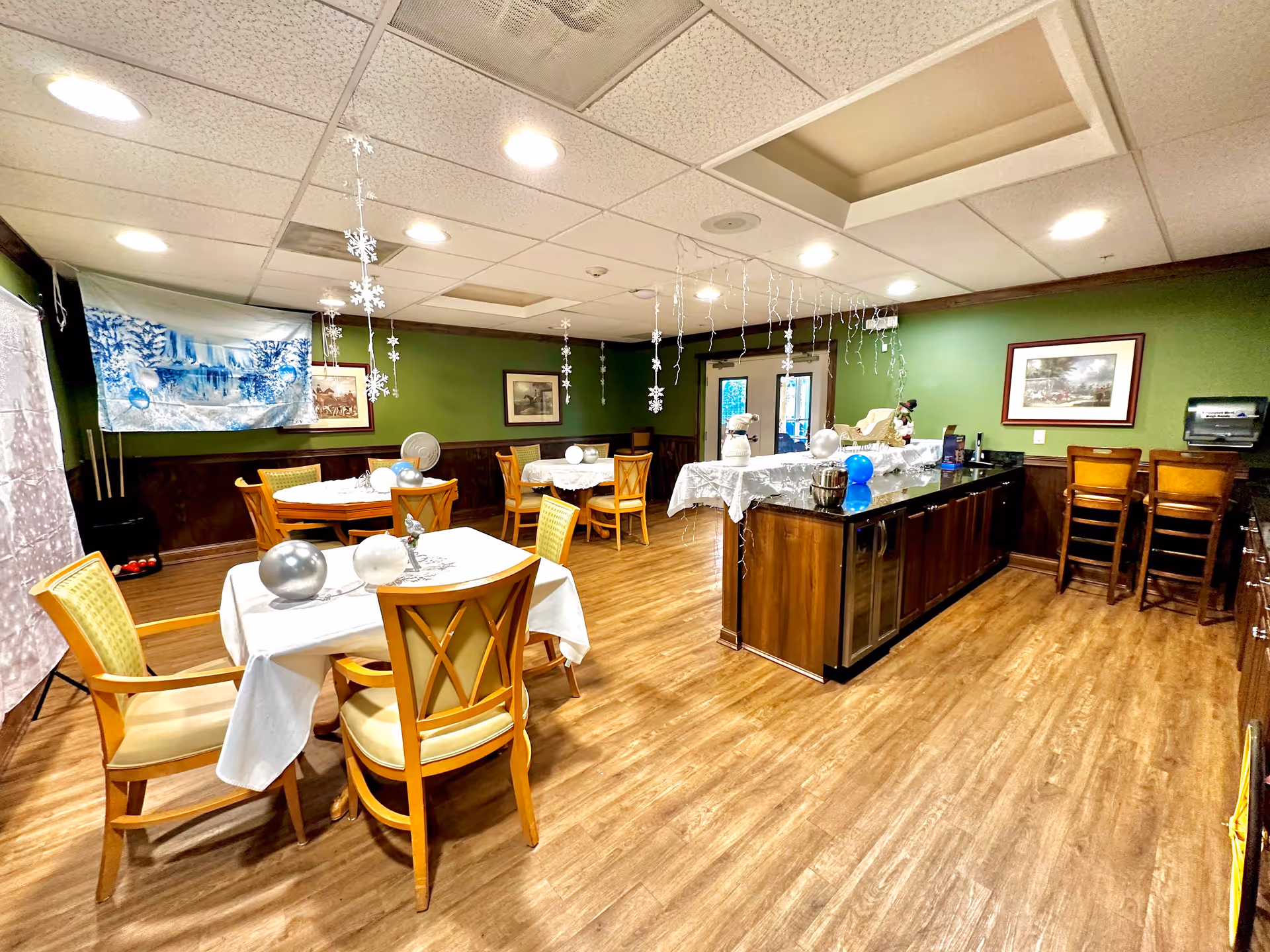 Bright dining/activity room with round tables and chairs, a central wood island, green walls, hanging snowflake decorations, and wood flooring.