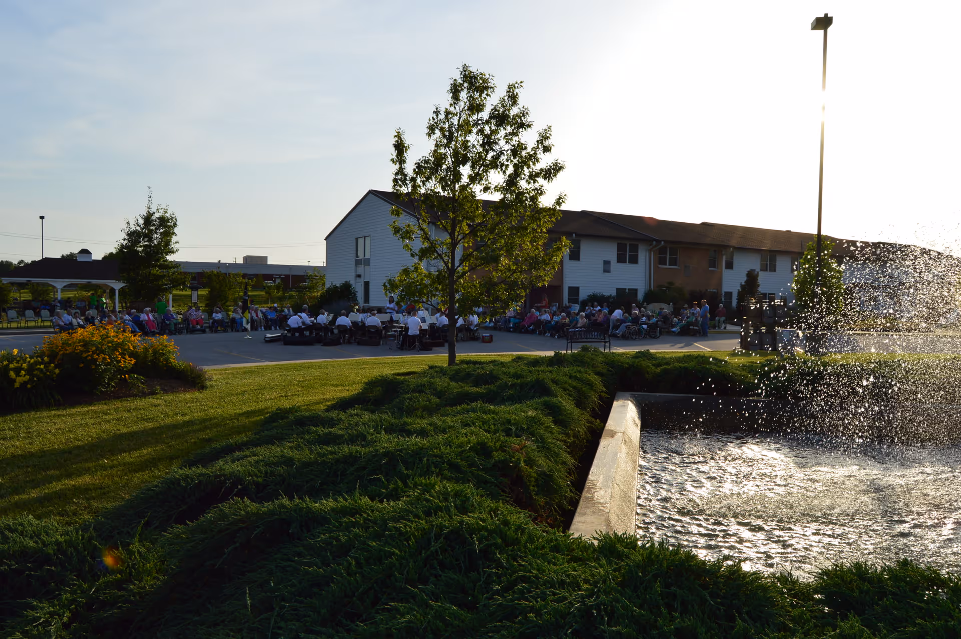 Outdoor scene at Magnolia Terrace Senior Living Apartments showing a group of people seated in chairs in a courtyard area near a two-story building. There is a water fountain spraying water on the right side, green grass, bushes, and a tree in the foreground under a clear sky.