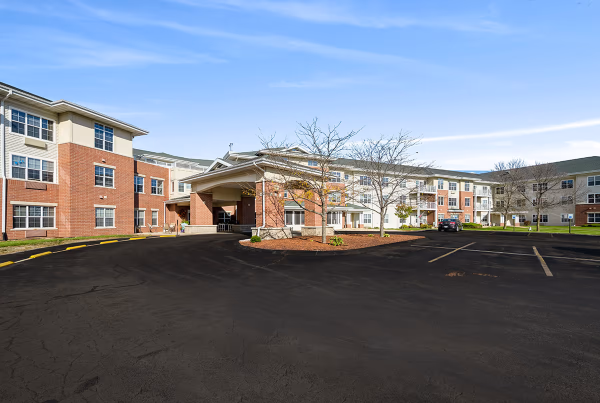 Exterior view of a senior living facility named Trustwell Living at Eagle Pointe Place, showing a large three-story building with brick and beige siding, multiple windows, a covered entrance, a parking lot with a few cars, and some leafless trees under a clear blue sky.