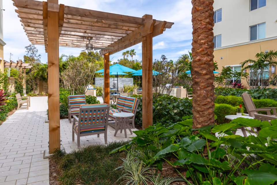 Outdoor seating area at The Club at Boynton Beach featuring a wooden pergola with ceiling fan, colorful cushioned chairs, small tables, lush green plants, palm trees, and blue umbrellas in the background under a partly cloudy sky.