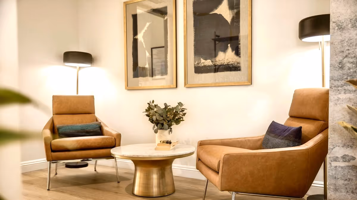 Two leather armchairs with pillows flank a round marble-top coffee table and floor lamps in a cozy seating area with framed art on the wall.
