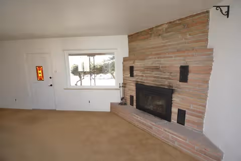 Empty living room with a stone fireplace, a large front window, and an entry door.