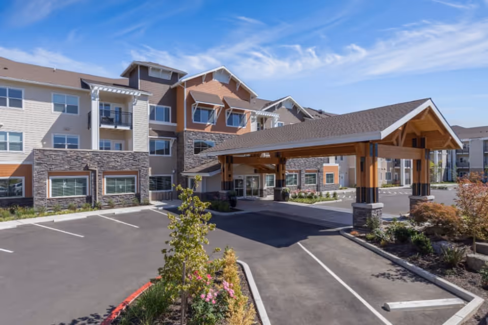 Front entrance of a senior living building with a covered porte-cochere, landscaped beds, and adjacent parking.