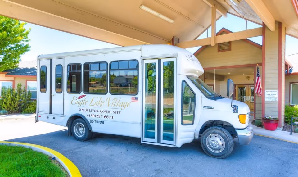 A white Eagle Lake Village senior living community shuttle bus parked under the covered entrance of the building.