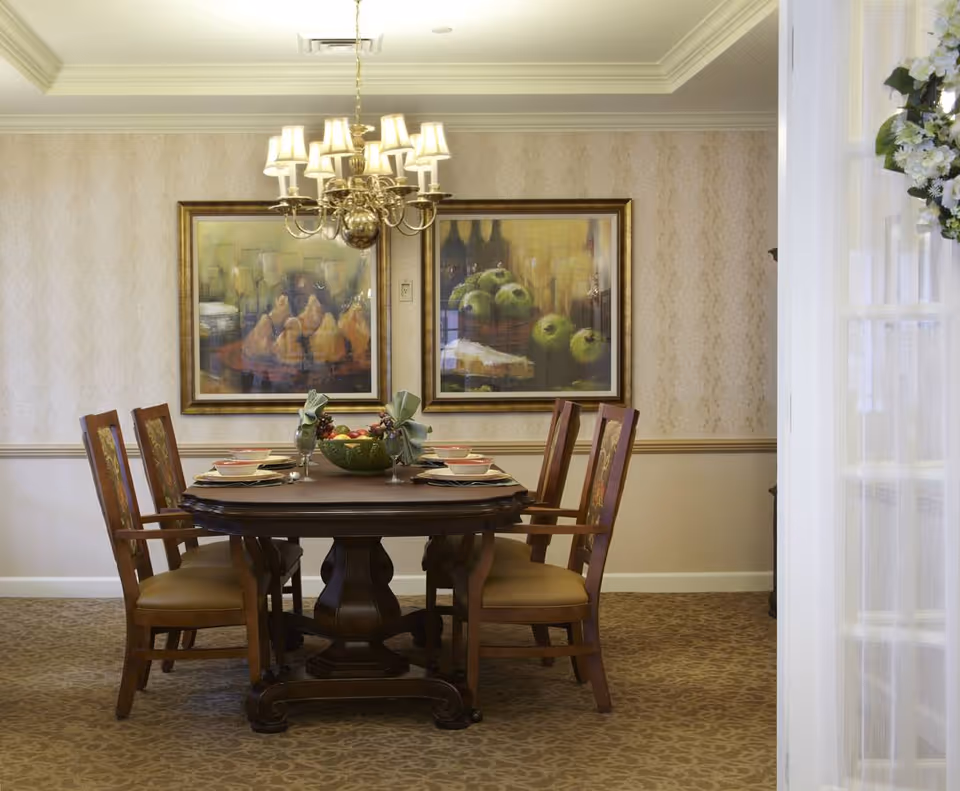 A formal dining room with a dark wooden table set for four, featuring plates, bowls, and napkins folded in a decorative manner. The room has patterned wallpaper, a beige carpet, two framed paintings of fruit on the wall, and a brass chandelier with multiple lampshades hanging above the table. A floral wreath is partially visible on the right side near a doorway.