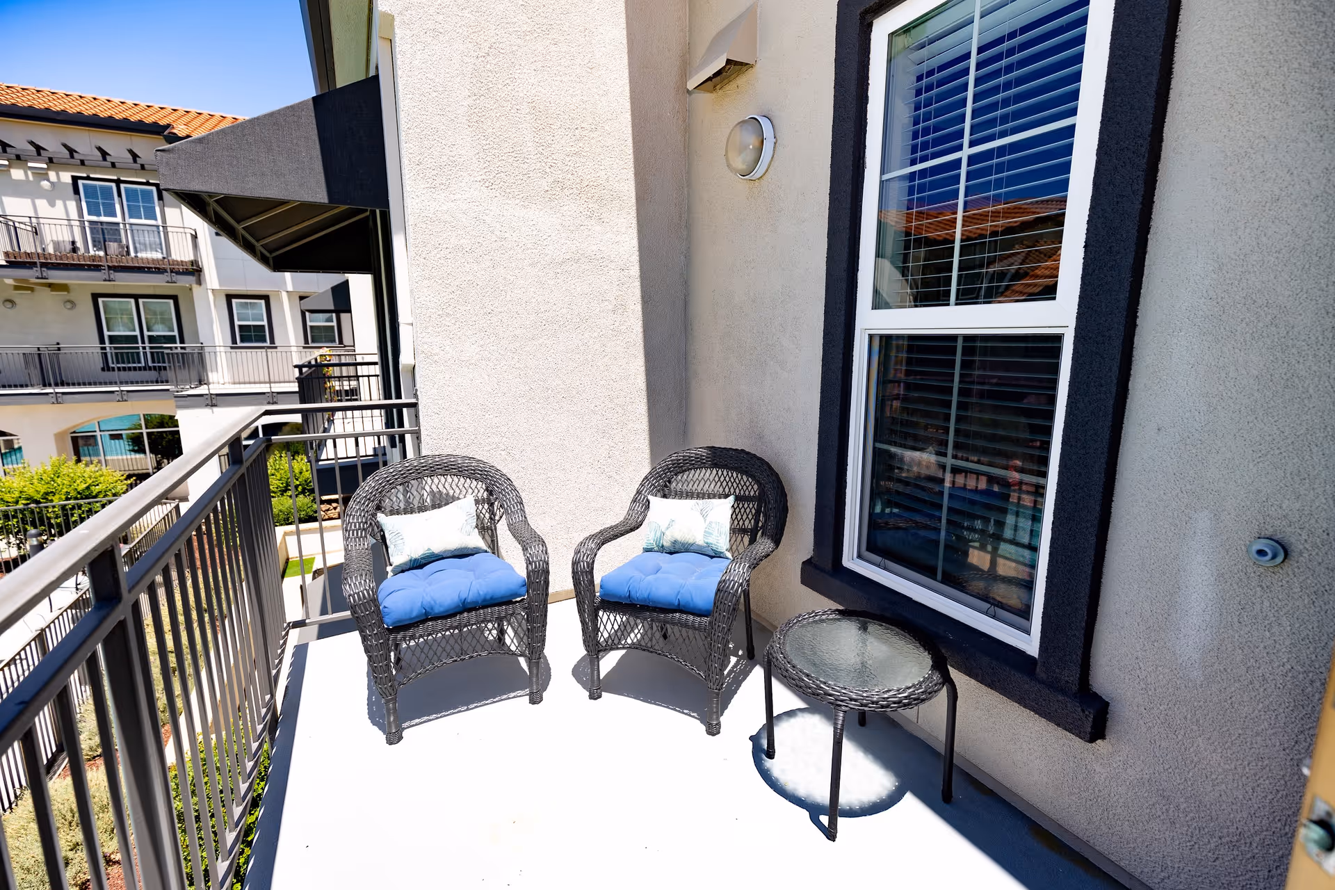 A small outdoor balcony area with two black wicker chairs with blue cushions and decorative pillows, and a small round glass-top table. The balcony has a metal railing and overlooks a courtyard with other apartment balconies visible in the background.
