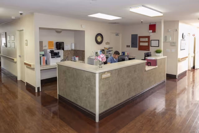 Reception desk and nurses' station in a senior care facility hallway with a staff member working at a computer.
