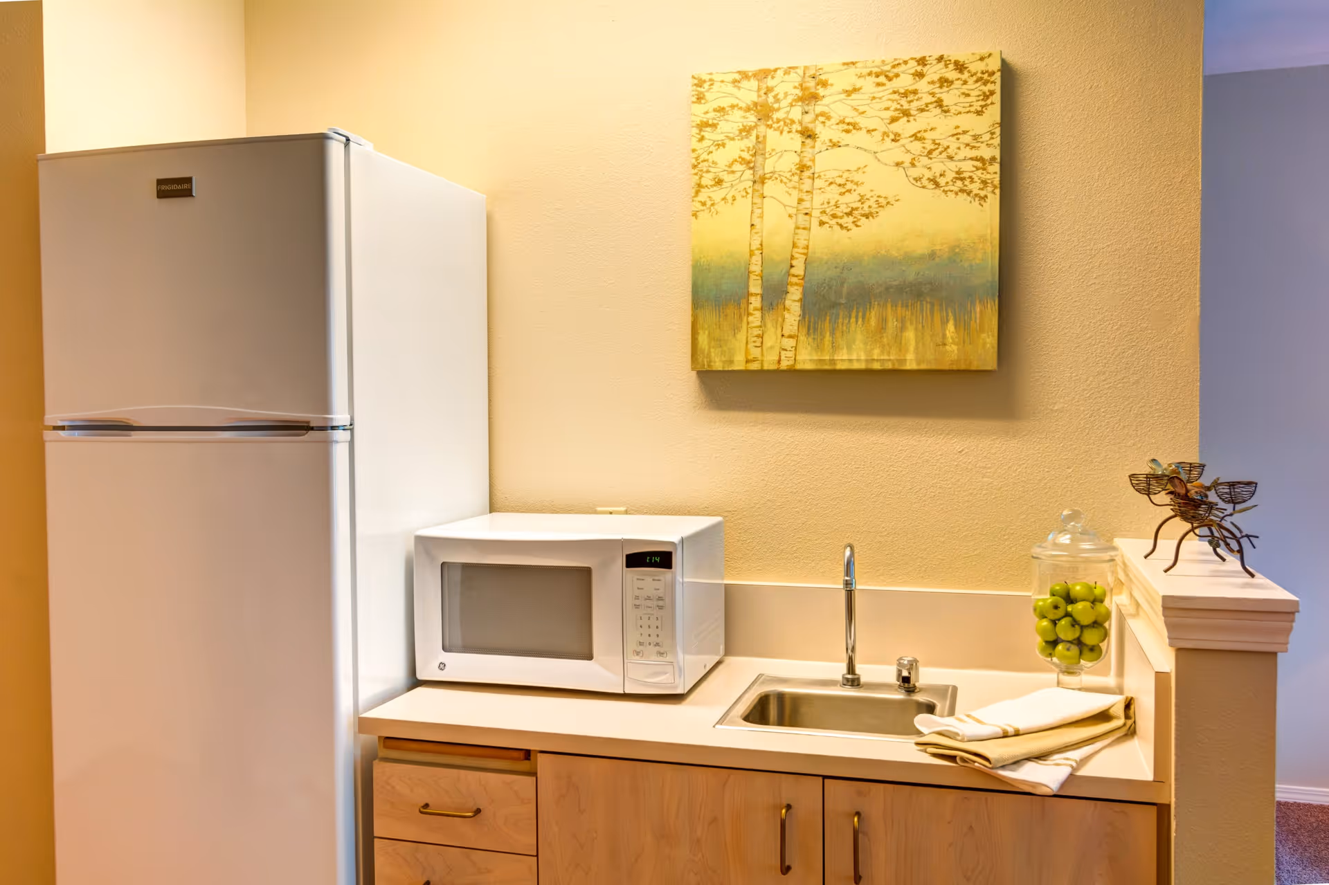 A small kitchenette area with a white refrigerator, a white microwave on the countertop, a stainless steel sink with a faucet, a glass jar filled with green apples, and a decorative metal butterfly sculpture. Above the countertop is a painting of two birch trees with a yellow and green background.
