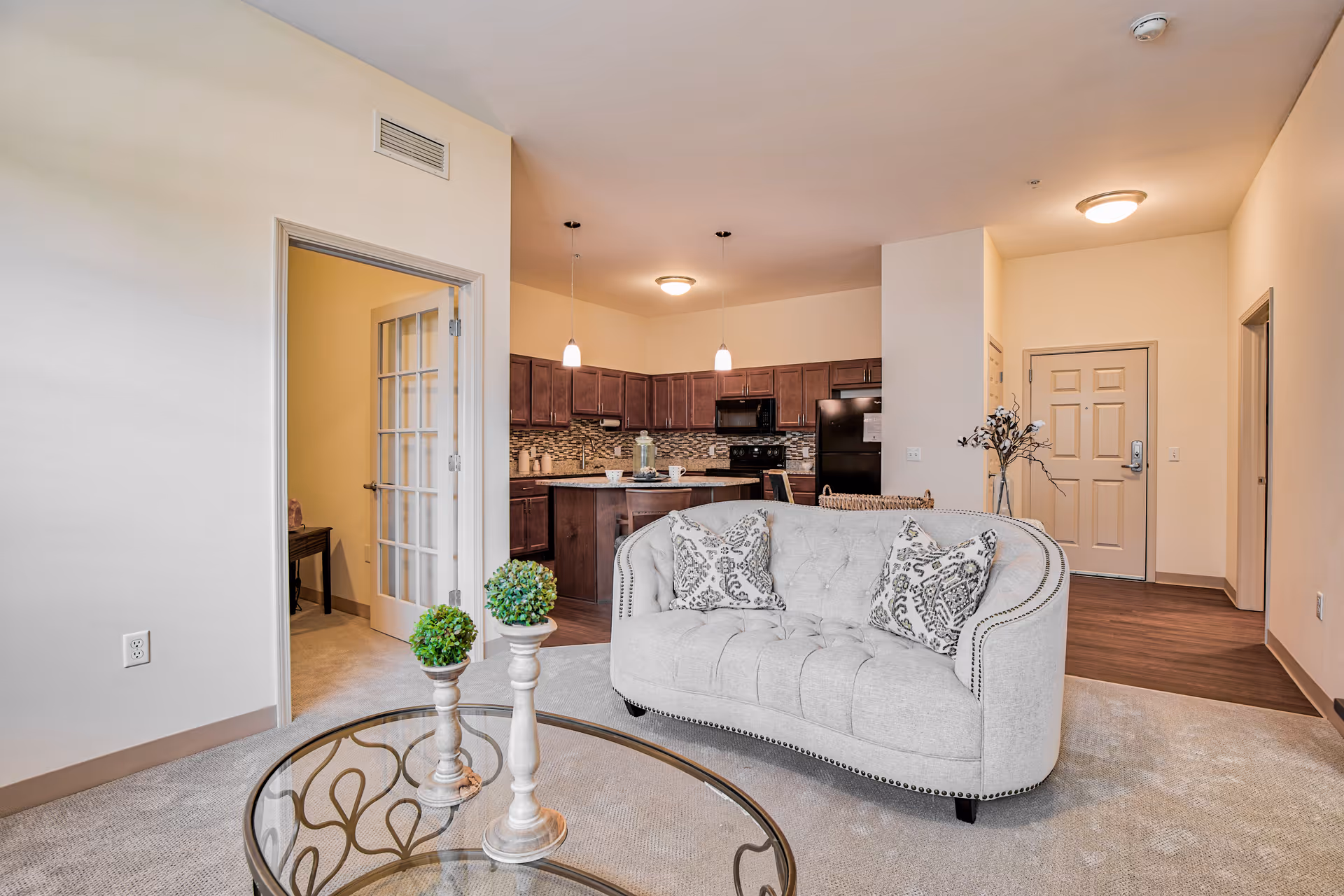 Interior view of a senior living facility apartment showing a living room with a light gray tufted loveseat with two patterned pillows, a glass coffee table with decorative plants, and an open kitchen with dark wood cabinets, black appliances, and pendant lights. There is a hallway with a closed door and an open door with glass panels leading to another room.