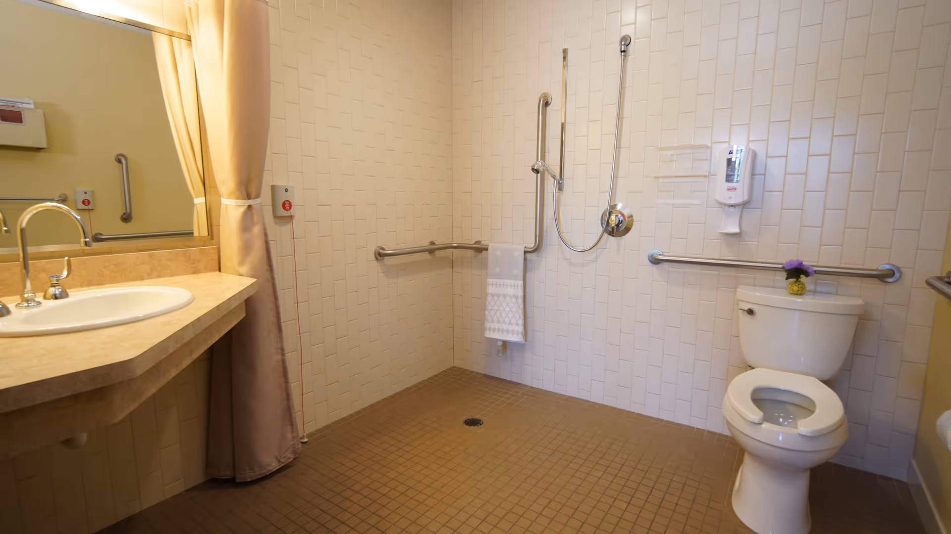 Accessible bathroom with beige tiled floor and white tiled walls. The room features a toilet with a grab bar on the wall behind it and to the side, a wall-mounted soap dispenser, a shower area with a handheld showerhead and grab bars, a towel hanging on one of the bars, a sink with a countertop, and a large mirror partially covered by a beige curtain.