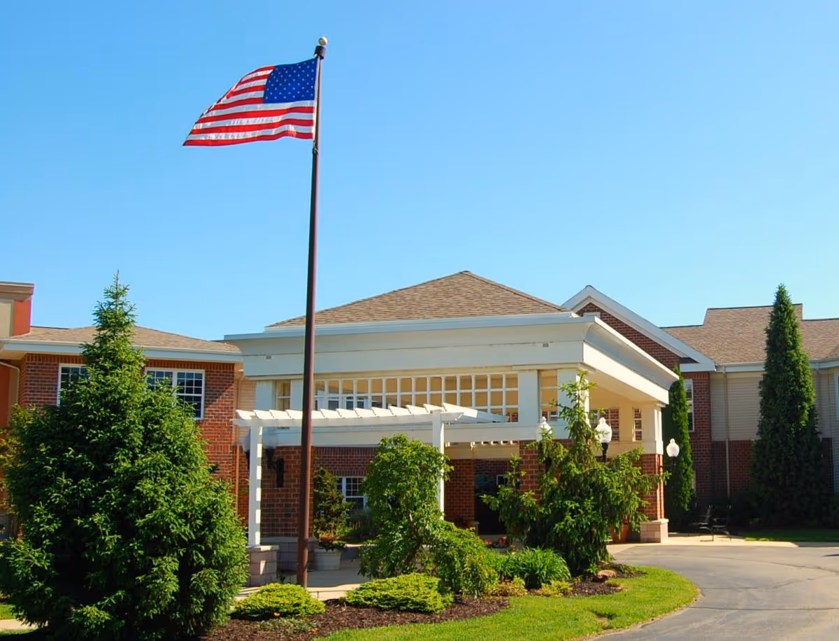 Exterior view of a senior living facility building with a covered entrance, surrounded by green bushes and trees, and an American flag flying on a flagpole against a clear blue sky.