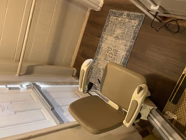A beige stairlift chair installed on a staircase with wooden flooring and a patterned rug at the bottom. The walls are light-colored with a handrail along the side. A white door with a push bar is visible at the bottom of the stairs.