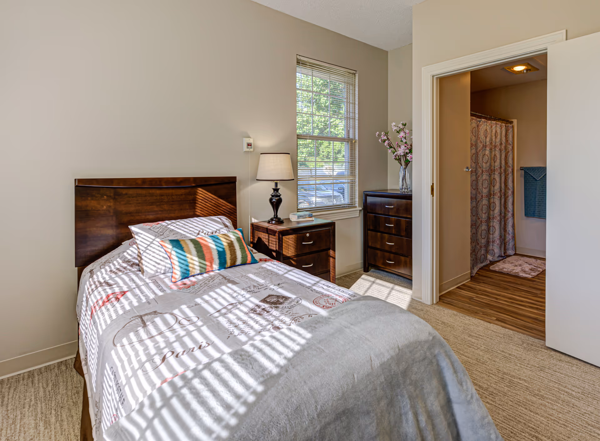 A bright bedroom with a single bed featuring a Paris-themed bedspread and colorful pillow. Next to the bed is a wooden nightstand with a lamp and a book. A window with blinds lets in natural light. There is a wooden dresser with a vase of flowers on top. An open door reveals a bathroom with a shower curtain and a towel hanging on the wall.