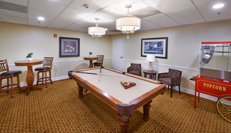 A recreational room with a pool table in the center, two chandeliers hanging from the ceiling, two round tables with chairs, two armchairs with a small table and lamp between them, framed pictures on the walls, and a red popcorn machine on the right side.