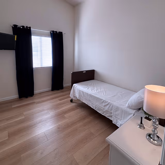 A simple bedroom with a single bed covered in white sheets, a bedside table with a lamp and a small plant, light wood flooring, a window with closed blinds and dark curtains, and a wall-mounted TV.