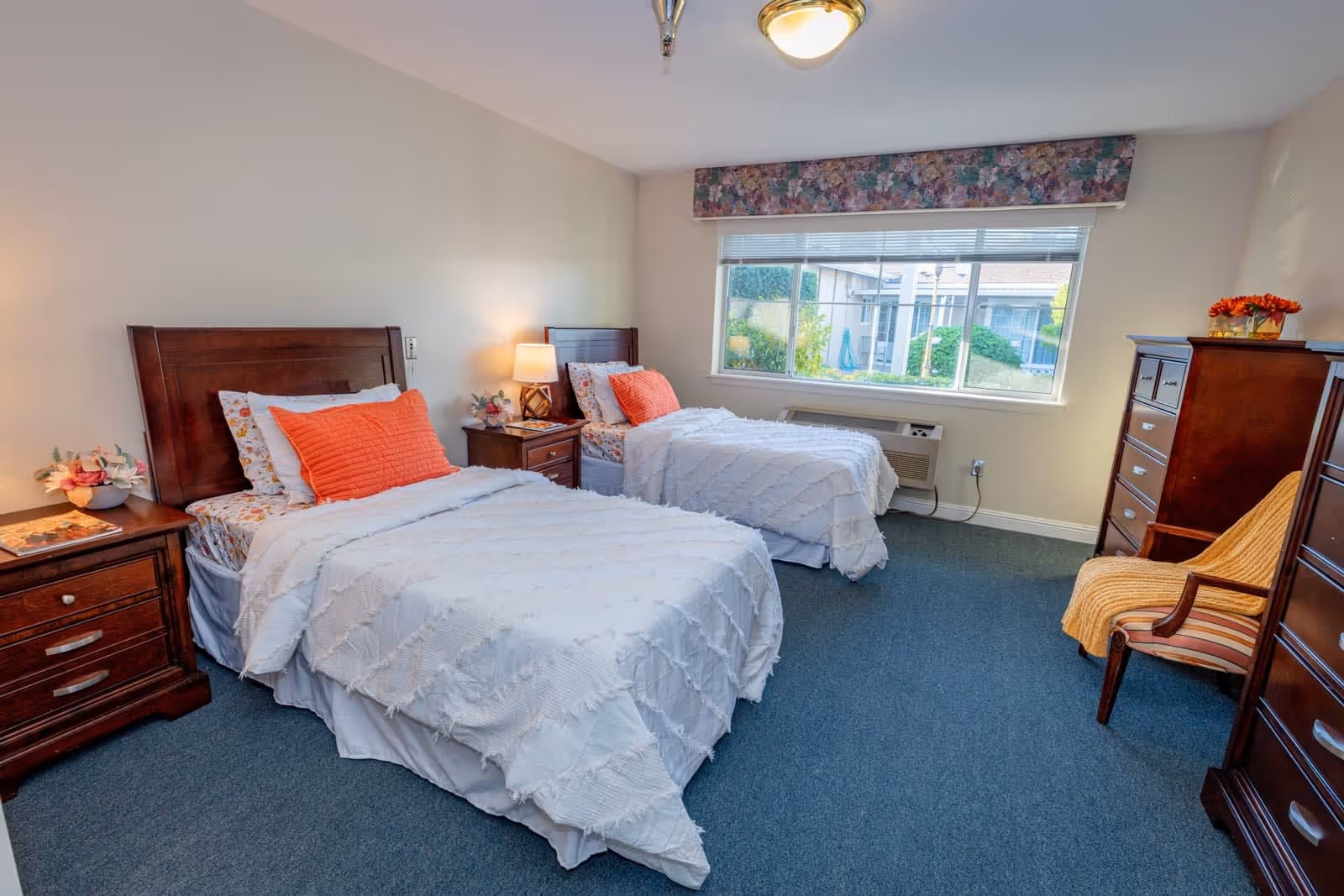 A bright and cozy bedroom with two twin beds featuring white textured bedspreads and orange accent pillows. Each bed has a wooden headboard and is flanked by wooden nightstands with lamps and floral decorations. A large window with floral valance lets in natural light, and a wooden dresser and chair with a yellow throw blanket are positioned on the right side of the room. The carpet is blue, and the walls are painted a light beige color.