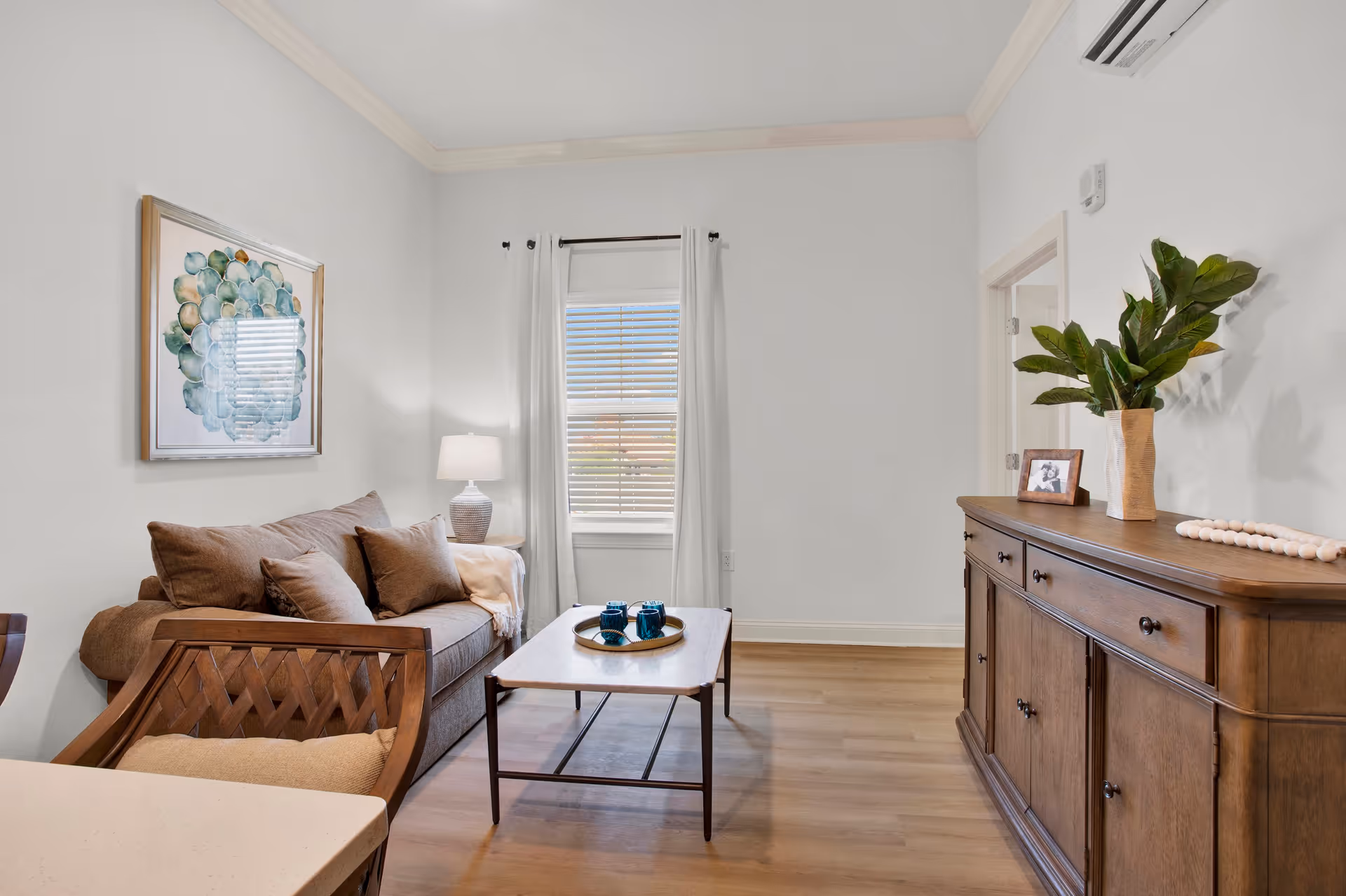 A cozy living room with a brown sofa adorned with cushions, a wooden coffee table with a tray holding blue cups, a wooden sideboard with a plant and a framed photo, a window with white curtains, and a lamp on a small side table next to the sofa.