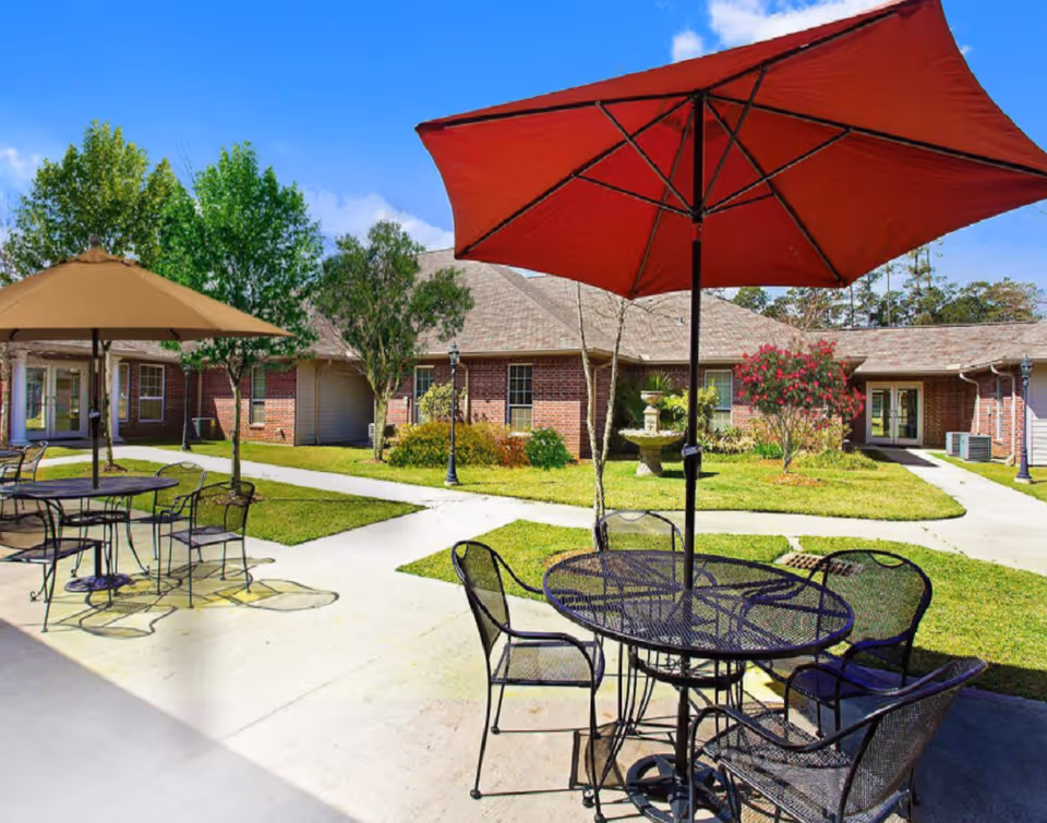 Outdoor patio area at Summerfield Senior Living with metal tables and chairs under large umbrellas, surrounded by green grass, trees, and a brick building under a blue sky.