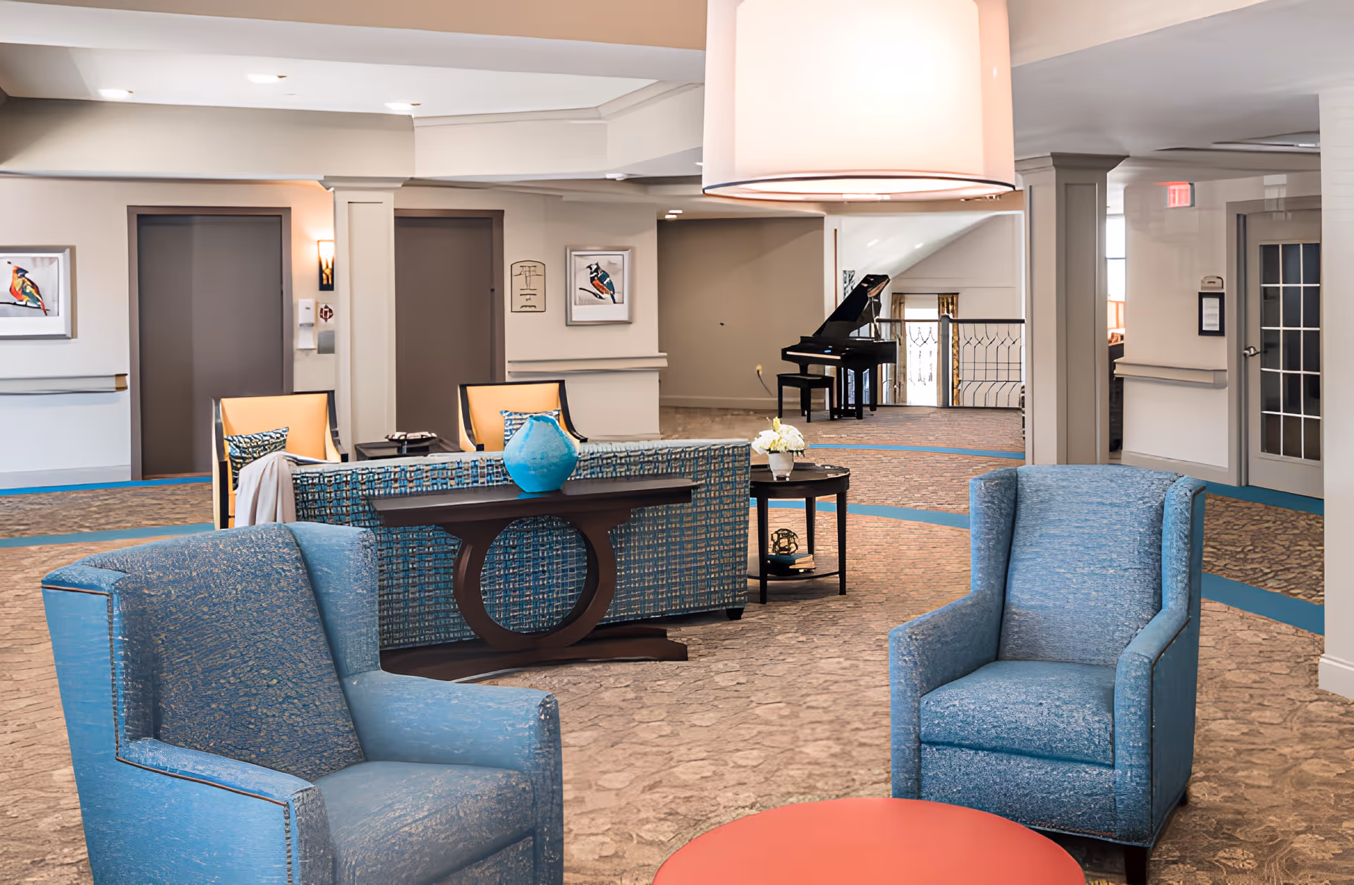 A spacious senior living facility common area with two blue armchairs in the foreground, a patterned sofa with a blue vase on a console table behind it, and a black grand piano in the background. The room has beige walls, carpeted floors, framed bird artwork, and soft lighting.