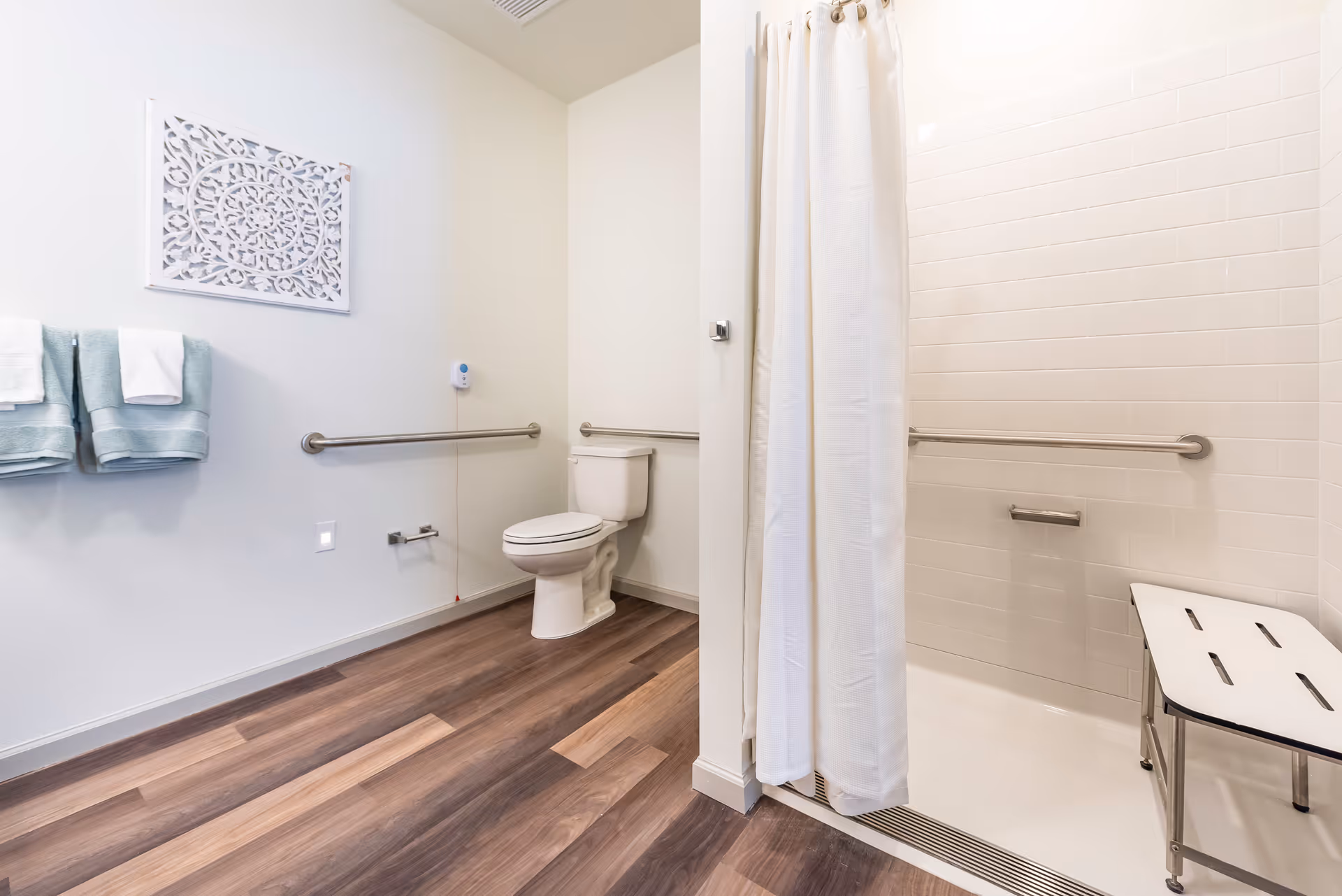 A clean and accessible bathroom with a white toilet, grab bars on the walls, a walk-in shower with a white curtain and a shower bench, light blue towels hanging on a rack, and decorative wall art. The floor has wood-style vinyl planks.