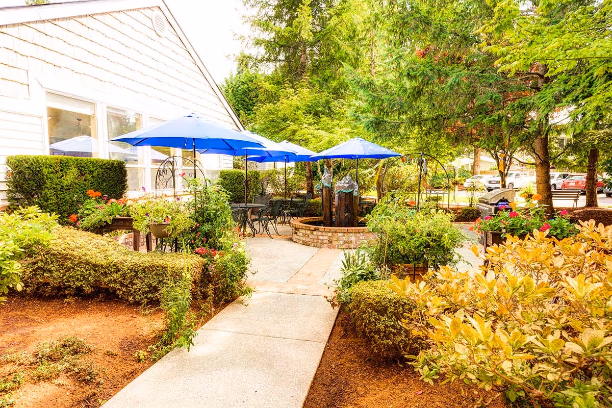 Outdoor patio area at The Gardens at Marysville featuring a paved walkway, neatly trimmed bushes, colorful flowers, several blue umbrellas shading metal tables and chairs, and a brick circular planter with a decorative water feature, surrounded by tall trees and greenery.
