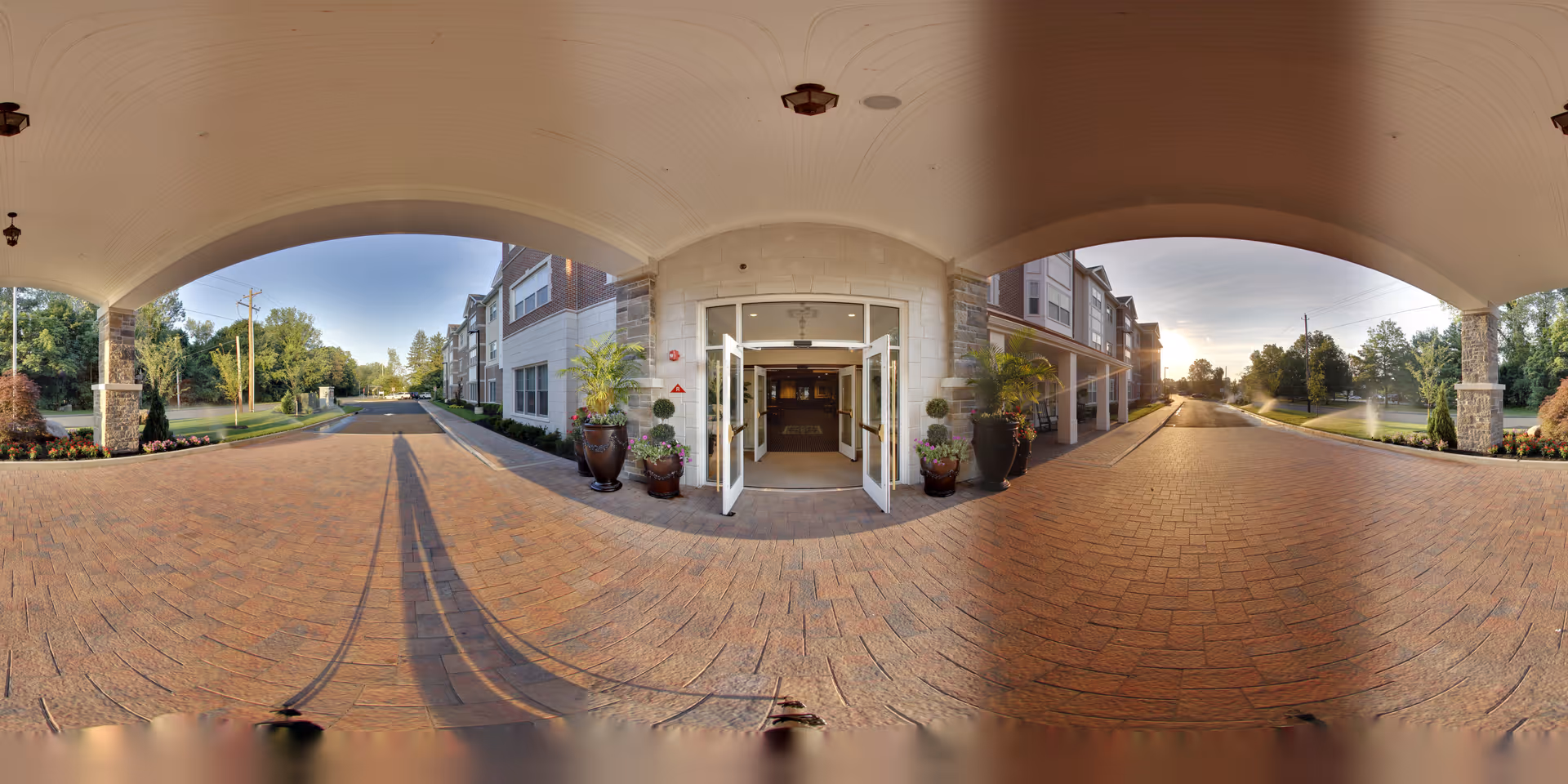 Covered entrance driveway of a senior living facility with open double glass doors leading inside. The area is paved with brick and flanked by large decorative planters with greenery. The building exterior features stone and brick accents, and there are trees and landscaped areas on either side of the driveway under a clear sky.