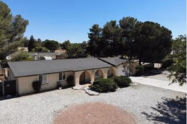 Single-story building with a dark roof and beige walls, featuring arched entryways and surrounded by trees and bushes under a clear blue sky. The foreground shows a gravel area with some landscaping.