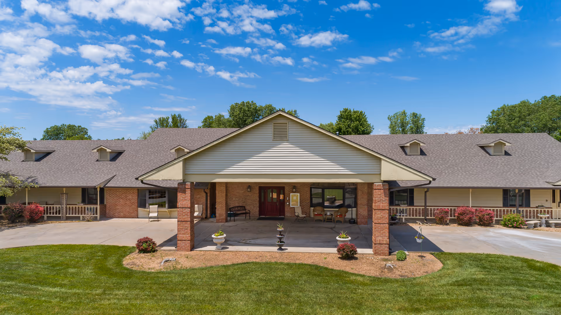 Front exterior view of a single-story senior living facility building with a covered entrance supported by brick pillars, surrounded by green lawn and landscaping under a partly cloudy blue sky.