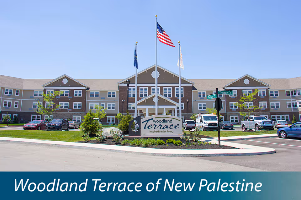 Front exterior view of Woodland Terrace of New Palestine senior living facility with a large building, three flagpoles displaying the American flag and two other flags, a landscaped area with a sign, and parked cars in the parking lot.