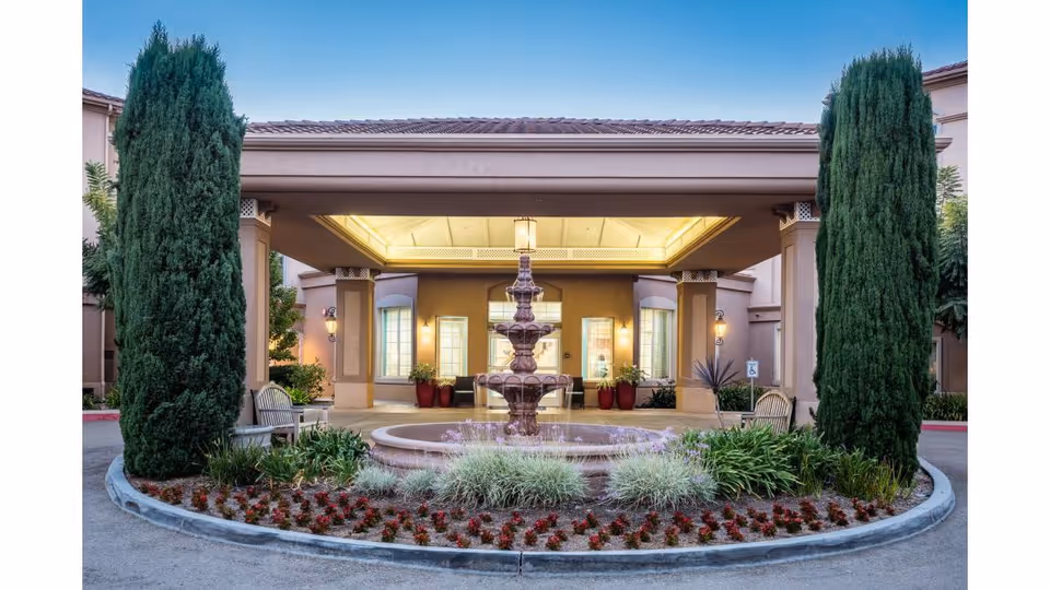 Entrance of Fairwinds - Ivey Ranch facility featuring a covered driveway with a central tiered water fountain surrounded by landscaped plants and flowers, flanked by two tall evergreen trees and benches on either side.
