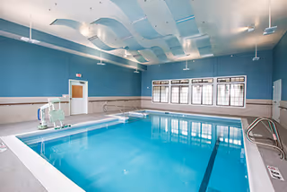 Indoor therapeutic swimming pool in a bright room with blue walls, ceiling acoustic panels, windows, and a pool lift.