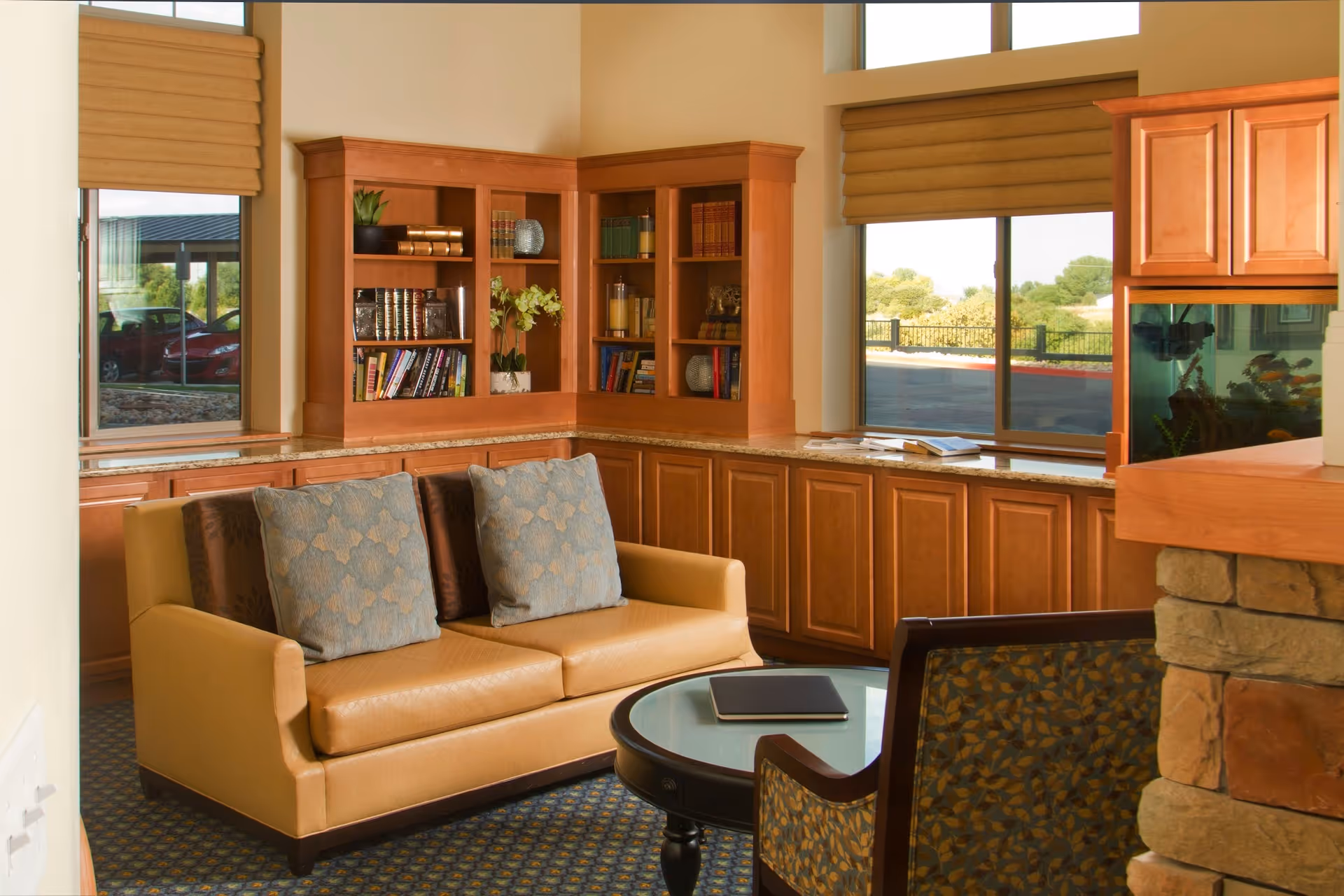 A cozy living room area with a tan leather sofa adorned with two blue patterned pillows, a round glass-top coffee table, and a patterned armchair. Behind the sofa is a built-in wooden bookshelf filled with books and decorative items. Large windows with brown blinds let in natural light and offer a view of the outdoors. The room features wooden cabinetry and a stone fireplace on the right side.