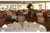 A spacious dining room with multiple round tables covered in white tablecloths, each set with napkins, glasses, and small flower arrangements. The room has large windows allowing natural light to fill the space, with chairs featuring various upholstery patterns arranged around the tables.